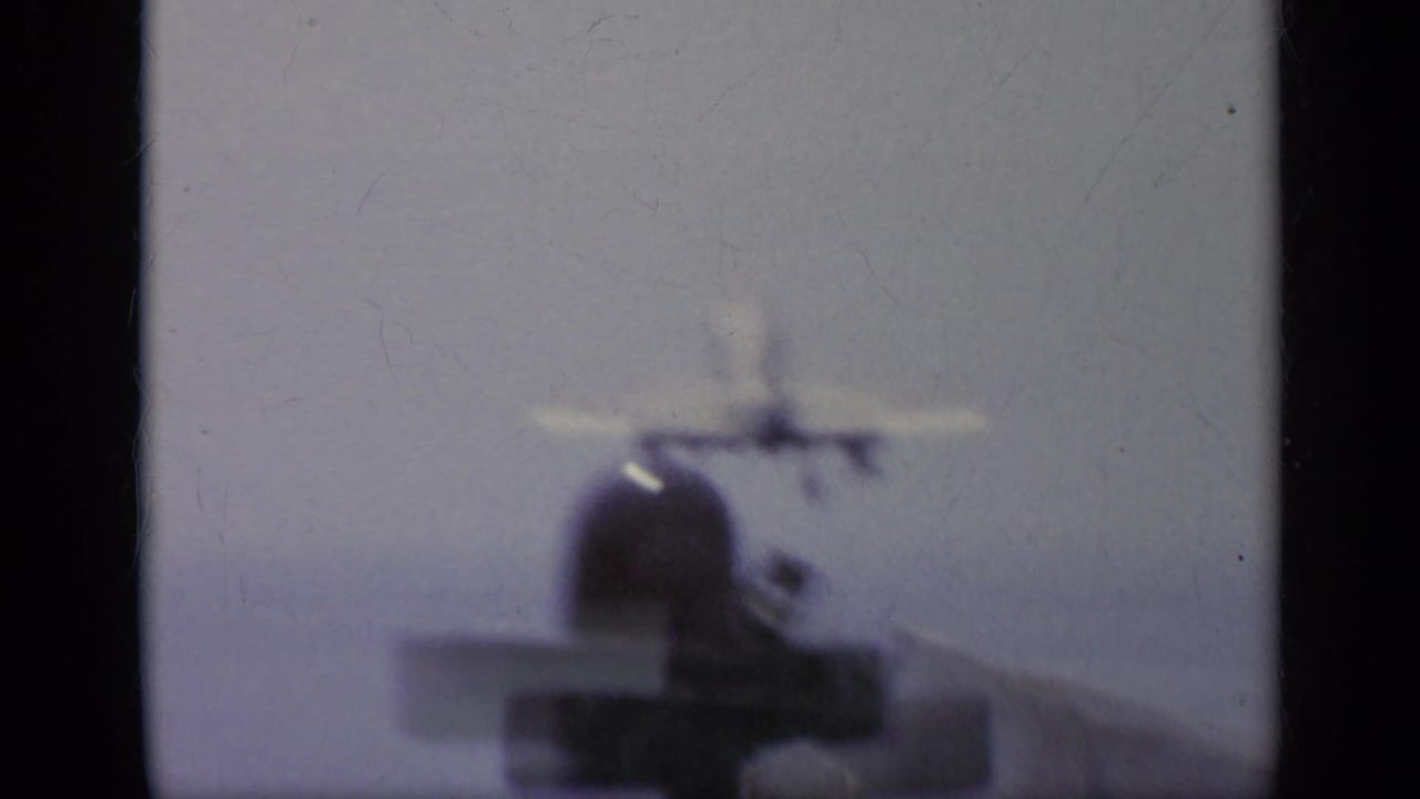 Soft 8mm view of a white jet on or near the carrier deck, with the aircraft blurred against a pale sea and sky background.