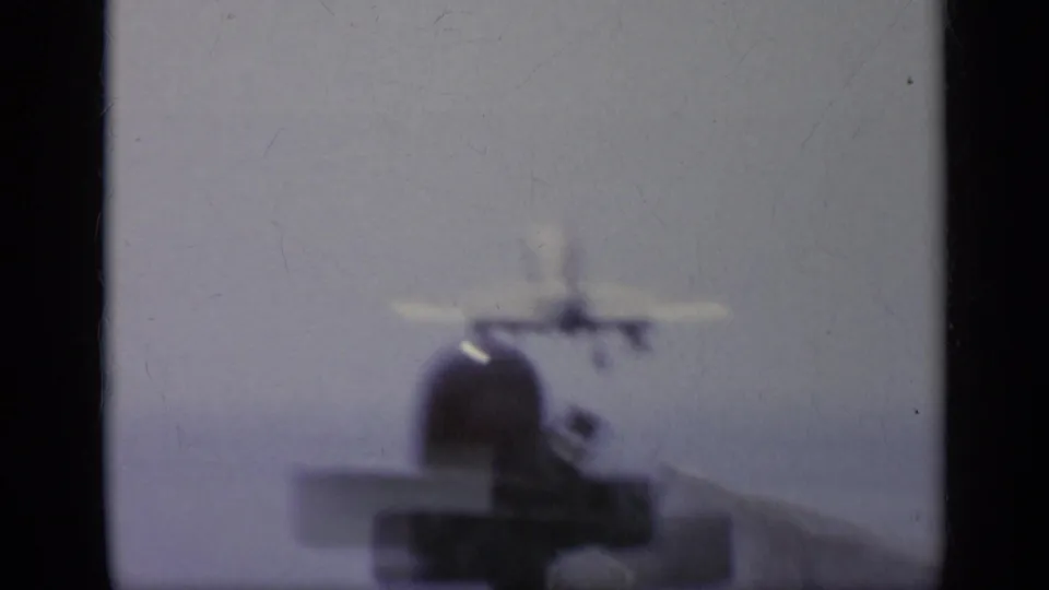 Soft 8mm view of a white jet on or near the carrier deck, with the aircraft blurred against a pale sea and sky background.