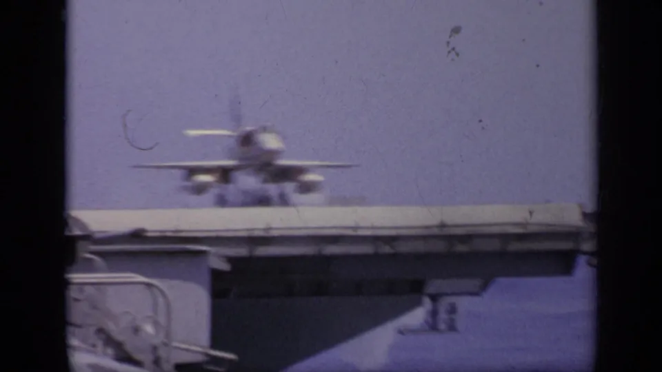 A military jet is seen near the carrier deck in archival 8mm footage, with the deck edge crossing the lower frame.