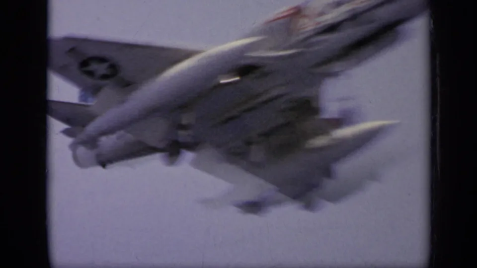 A military jet lifts off from the aircraft carrier deck, its tail and exhaust blurred by motion in the 8mm frame.