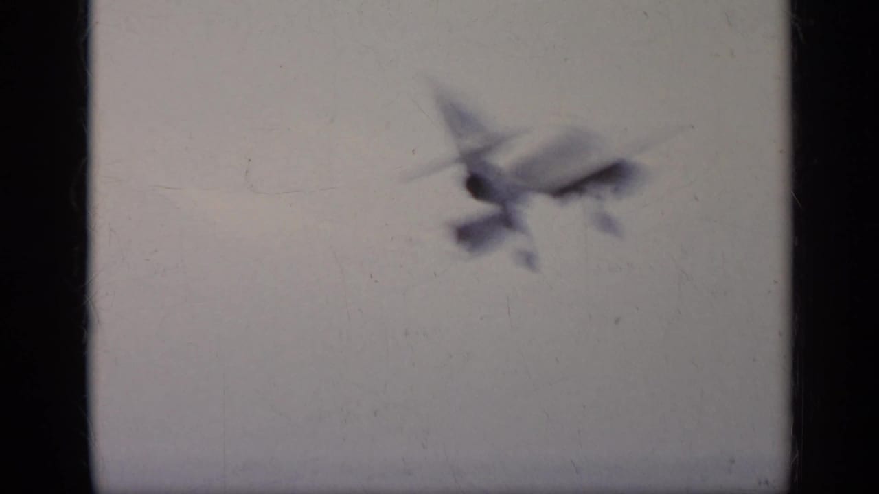 Soft 1962 8mm frame of an aircraft in flight against a pale sky, with visible film gate edges and grain.