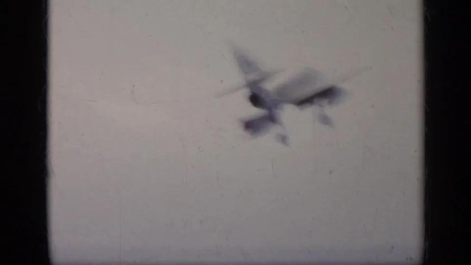 Soft 1962 8mm frame of an aircraft in flight against a pale sky, with visible film gate edges and grain.