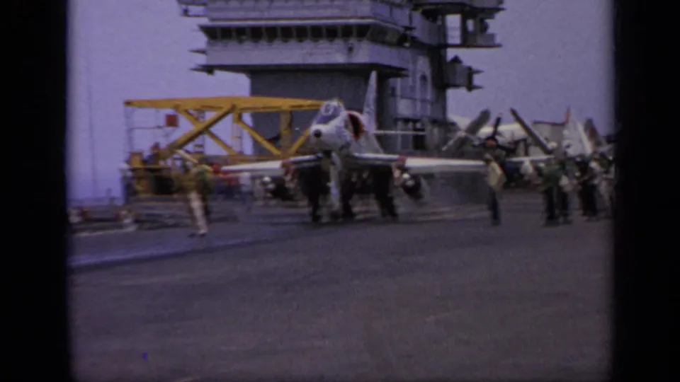 A white jet sits on an aircraft carrier deck in 1962 Italy footage, with crew nearby and the ship island behind it.