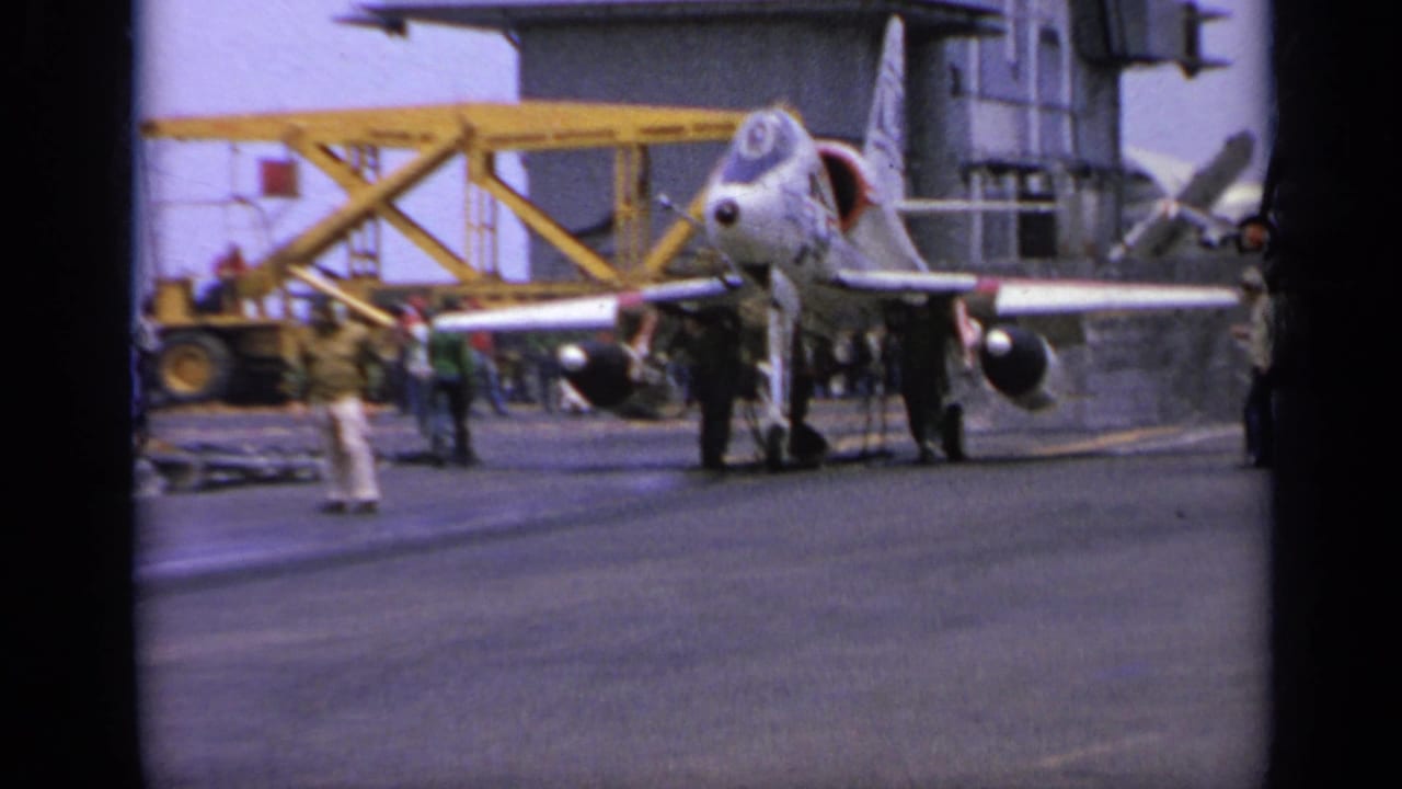 Military jet on an aircraft carrier deck with crew nearby and yellow deck equipment behind it.