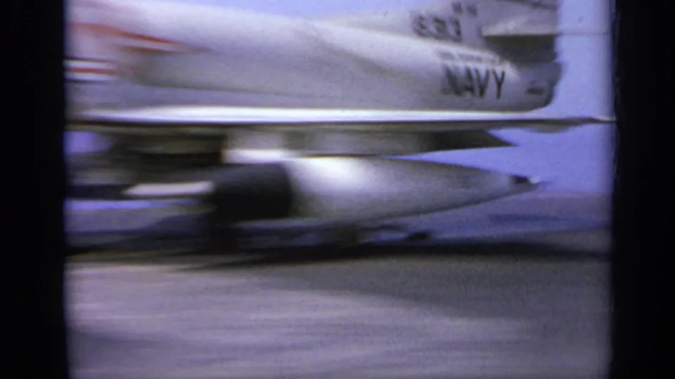 Motion-blurred 1962 home-movie frame of a Navy-marked jet accelerating across an aircraft carrier deck.