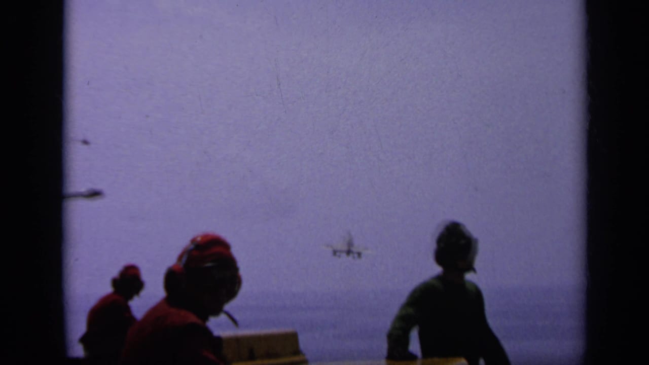 Servicemen watch a small aircraft approach over the sea from a ship deck.
