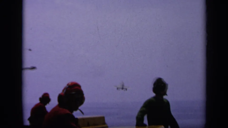 1962 Italy 8mm frame showing servicemen in headset gear watching a small aircraft approach over open water from a vessel.