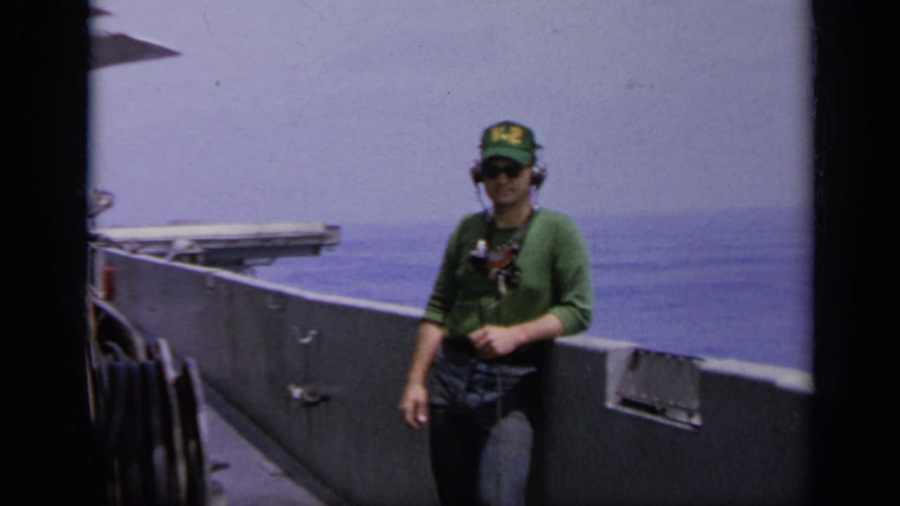 Man in green V-2 cap and headphones stands beside a ship rail at sea.