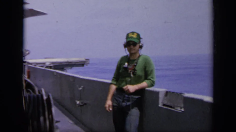 1962 Italy 8mm frame of a man in a green sweater, V-2 cap, and headphones standing on a vessel deck with ocean behind him.