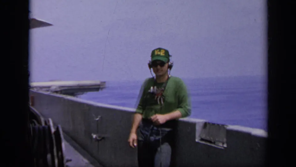 1962 Italy 8mm frame showing a man in a V-2 cap and headset walking near the rail of a vessel at sea.