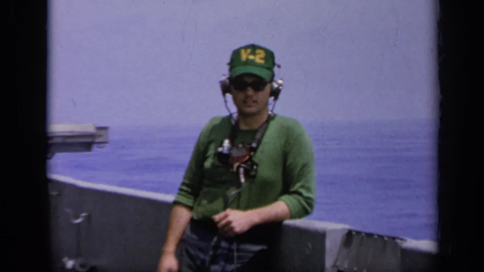 1962 Italy 8mm frame with a closer view of a headset-wearing man in a V-2 cap on a vessel, with calm sea in the background.