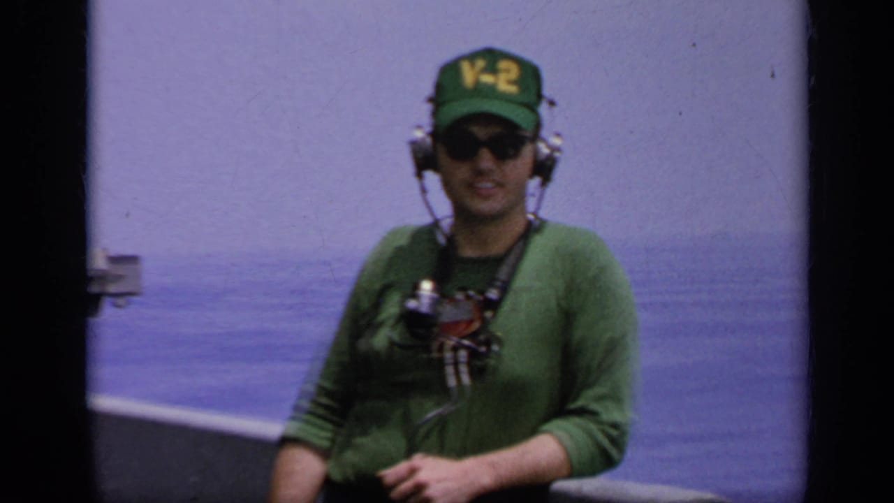 1962 Italy home-movie frame of a Navy crew member in a green V-2 cap, sunglasses, and headset on a ship deck at sea.