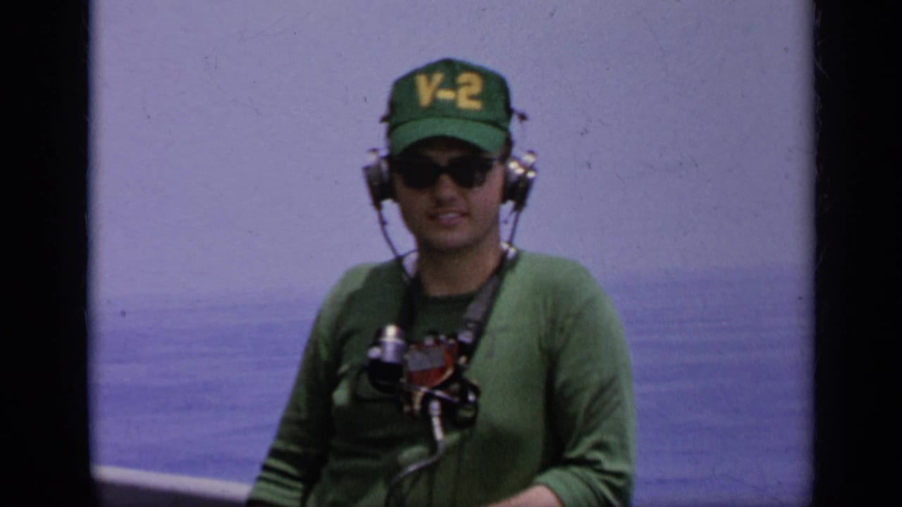 Navy crew member wearing a green V-2 cap, sunglasses, headset, and chest gear stands on deck in 1962 Italy footage.