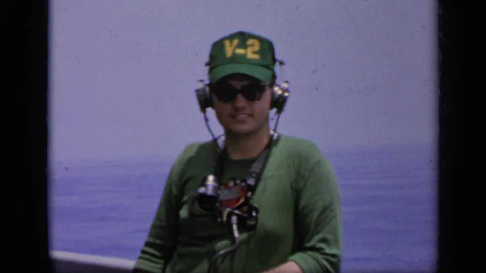 Navy crew member wearing a green V-2 cap, sunglasses, headset, and chest gear stands on deck in 1962 Italy footage.