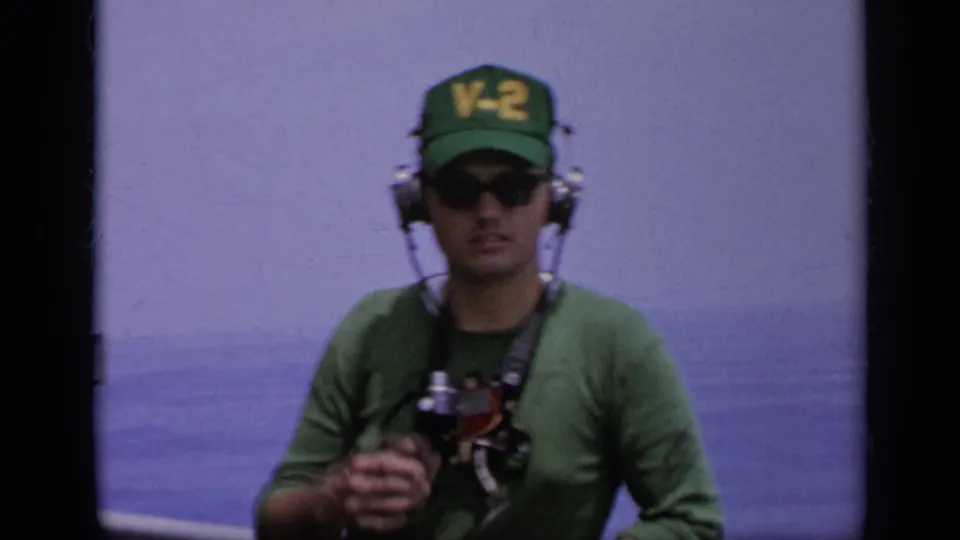 1962 8mm frame showing a Navy crew member in a green V-2 cap and headset holding gear on a ship deck at sea.