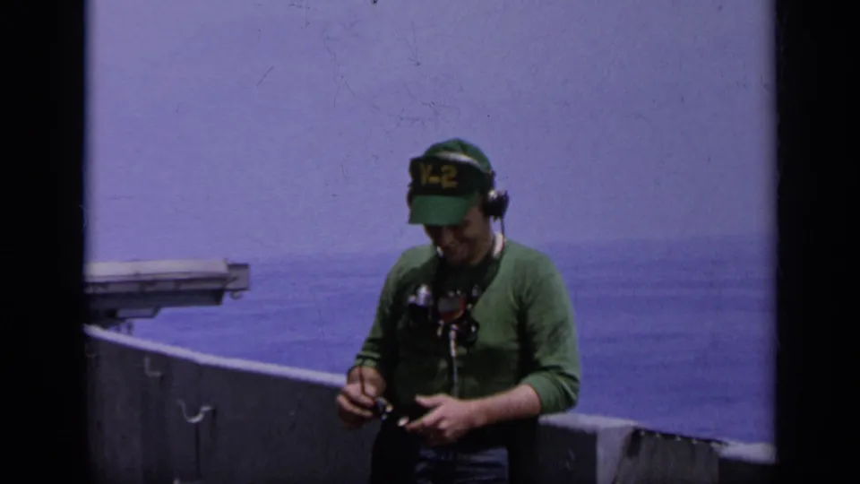 Navy crew member in green V-2 cap looks down at handheld gear on a ship deck in 1962 Italy home-movie footage.