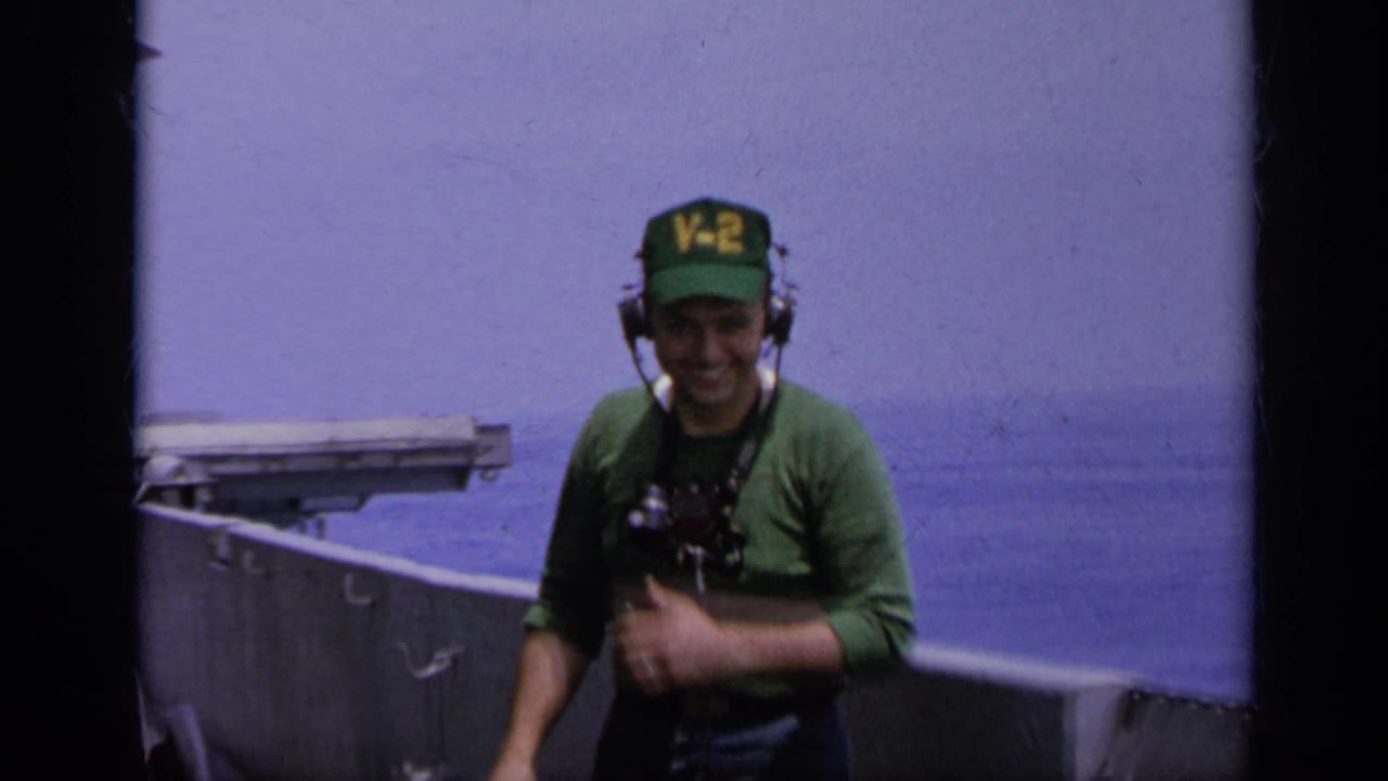Crewman in green V-2 cap and headset smiles on an aircraft carrier deck with ocean behind him.