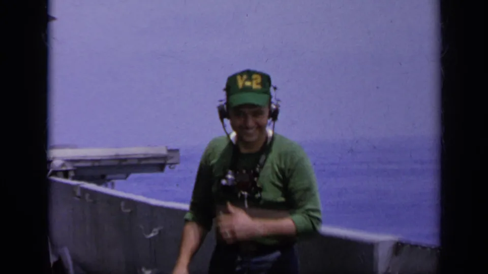 1962 Italy home-movie frame showing a U.S. Navy deck crewman in a green V-2 cap and headset smiling toward the camera near the carrier rail.