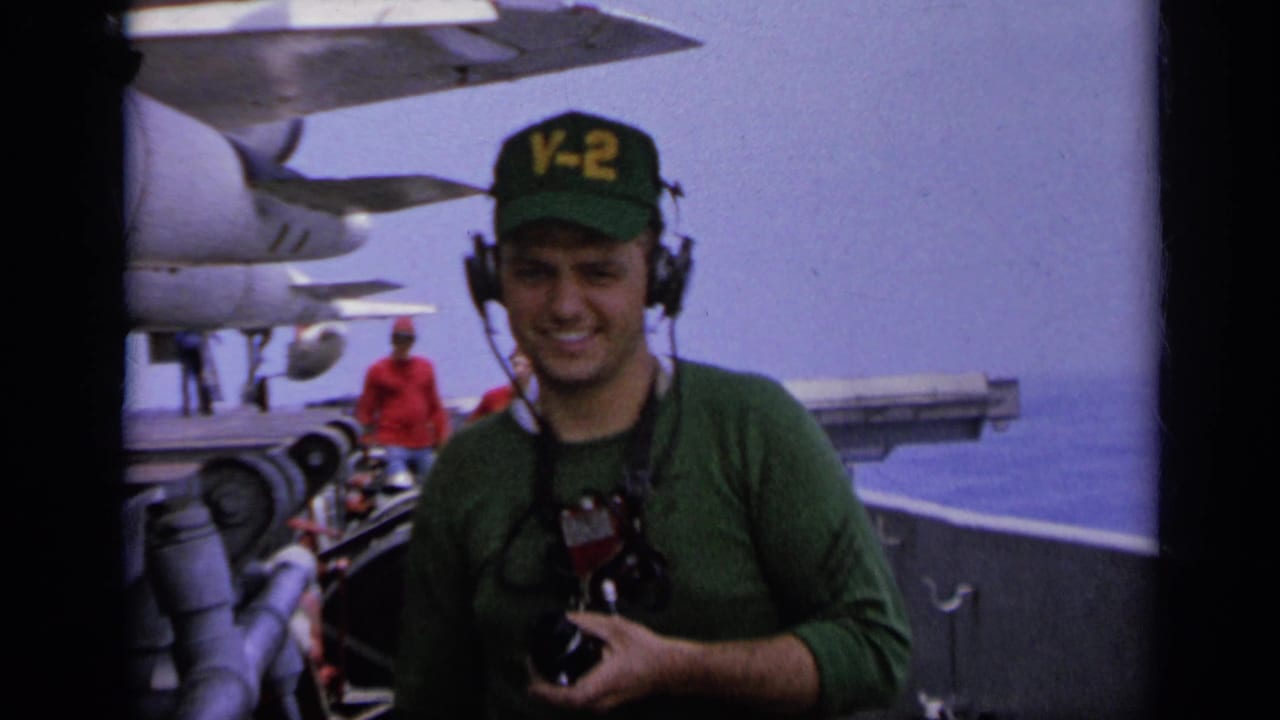 1962 Italy carrier-deck frame with a smiling U.S. Navy crewman holding a camera as aircraft and another deck figure appear in the background.