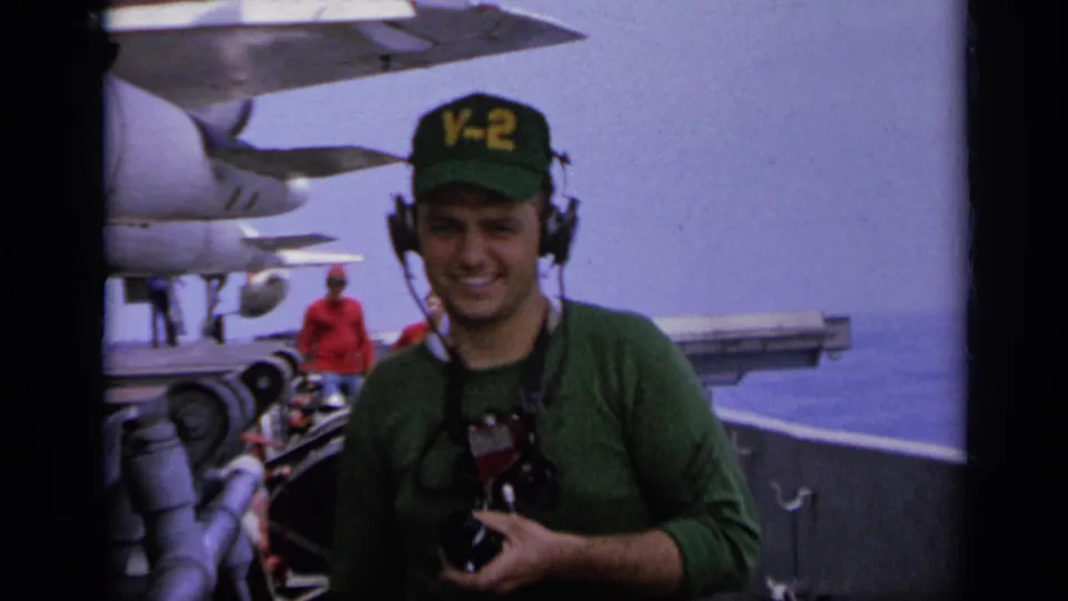 1962 Italy carrier-deck frame with a smiling U.S. Navy crewman holding a camera as aircraft and another deck figure appear in the background.