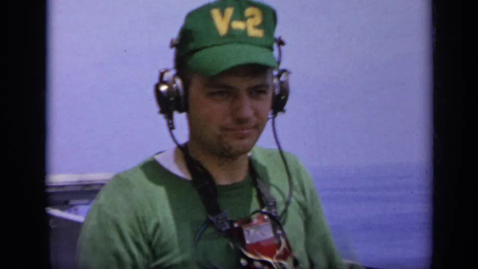 Close 1962 Italy home-movie view of a U.S. Navy deck crewman in green V-2 cap and headset, framed against the carrier edge and ocean.