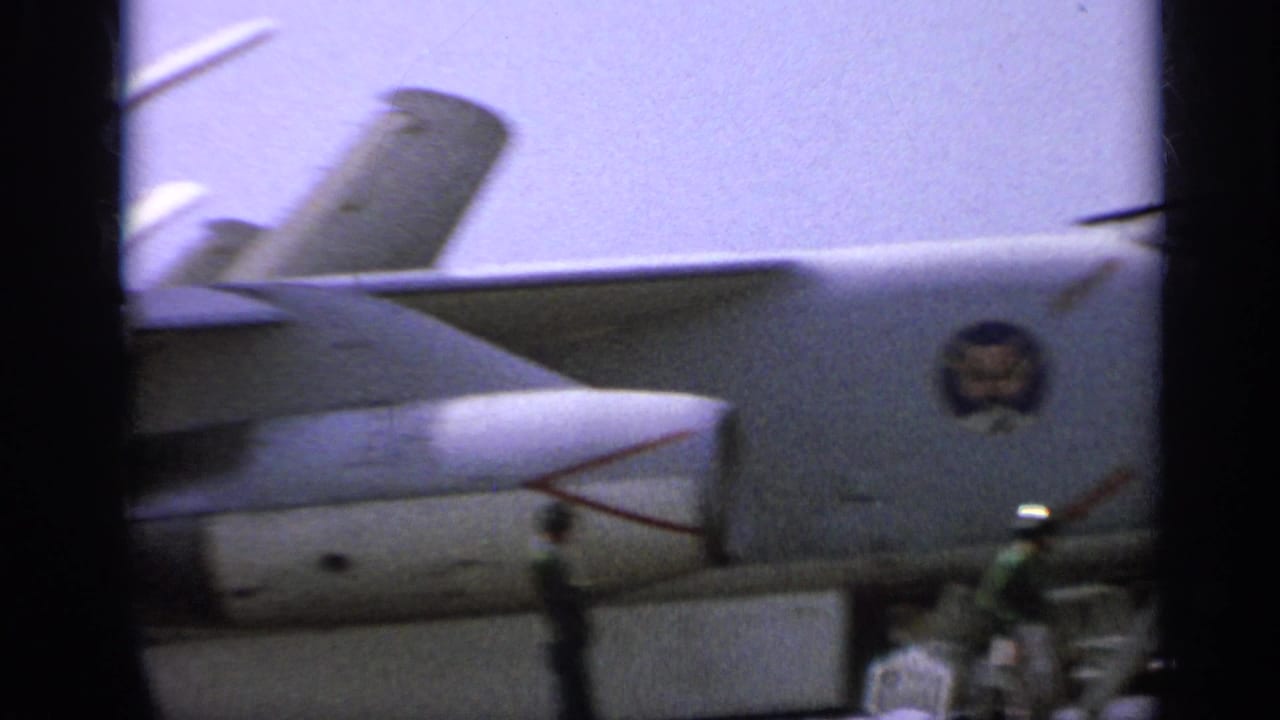 Close 1962 carrier-deck view of a U.S. Navy jet fuselage and tail, with military insignia and deck personnel in the background.