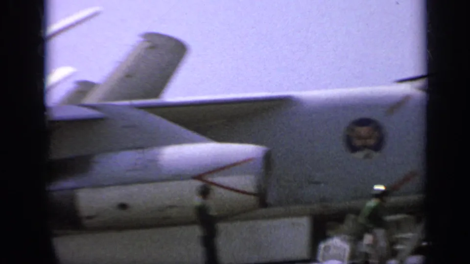 Close 1962 carrier-deck view of a U.S. Navy jet fuselage and tail, with military insignia and deck personnel in the background.
