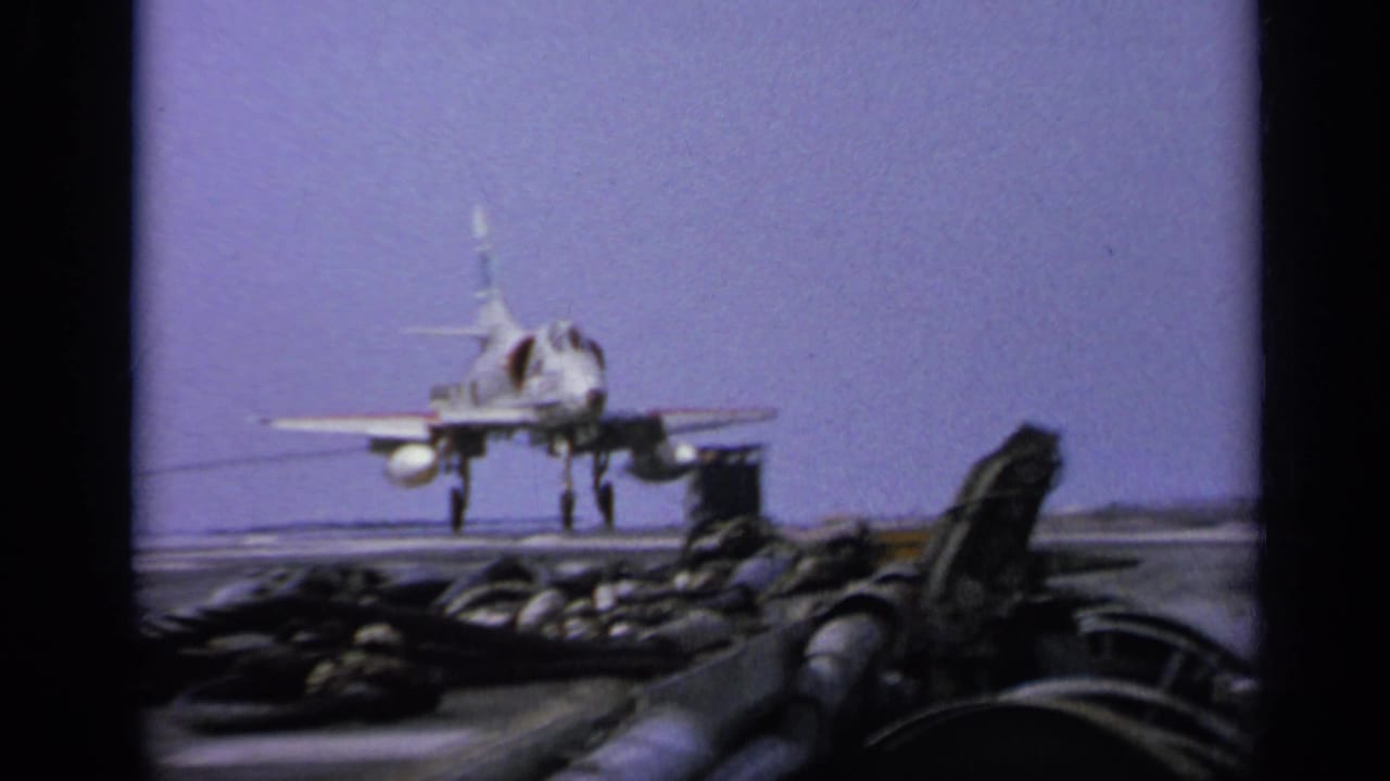 1962 8mm footage frame showing a jet fighter on an aircraft carrier deck in Italy, with deck equipment and pale sky visible.