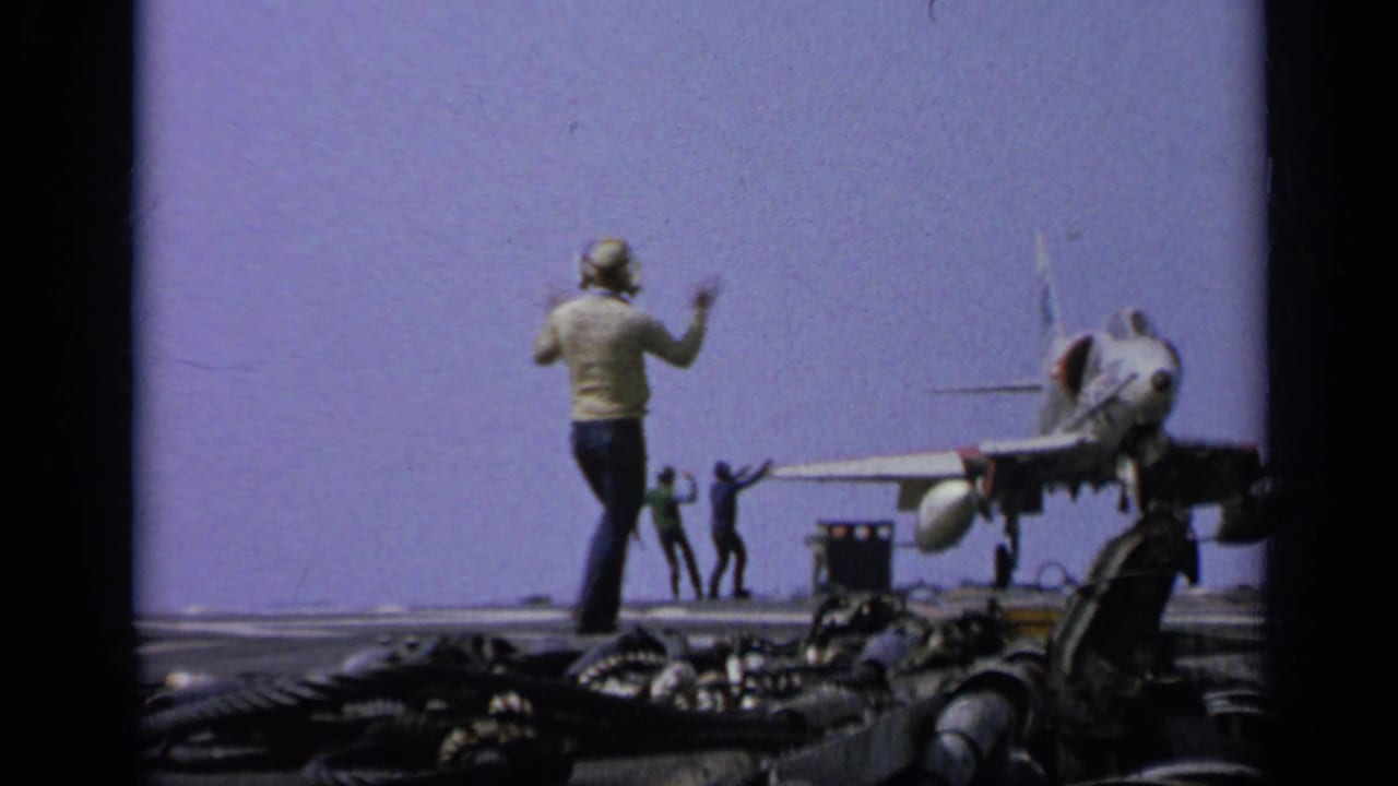 Crew member in helmet signals on the carrier deck as a jet fighter sits nearby, with other personnel visible in the distance.