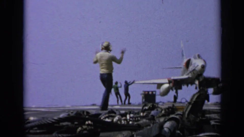 Crew member in helmet signals on the carrier deck as a jet fighter sits nearby, with other personnel visible in the distance.