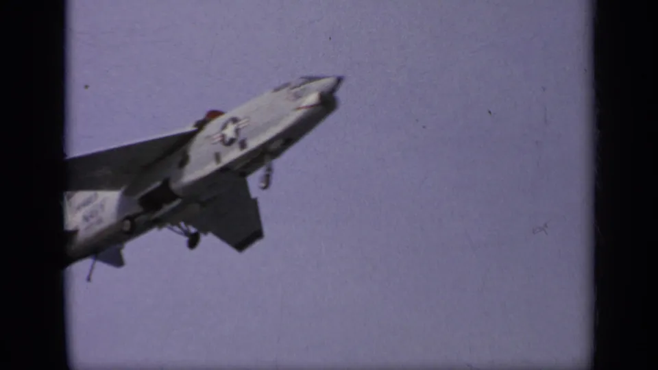 A close 8mm frame shows the jet fighter from below or alongside, with landing gear and military markings visible against the sky.