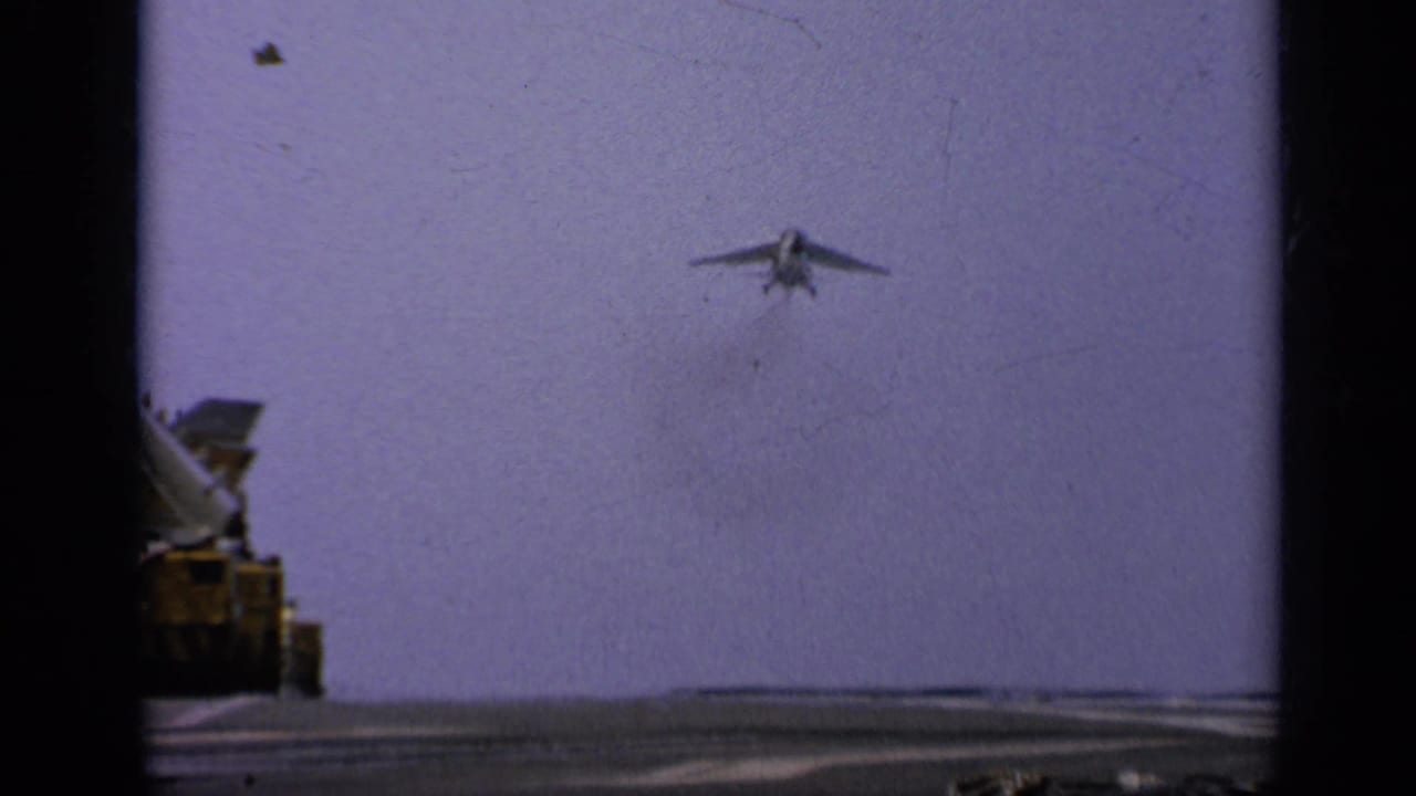 Jet approaches an aircraft carrier deck from a distance with deck equipment at left.