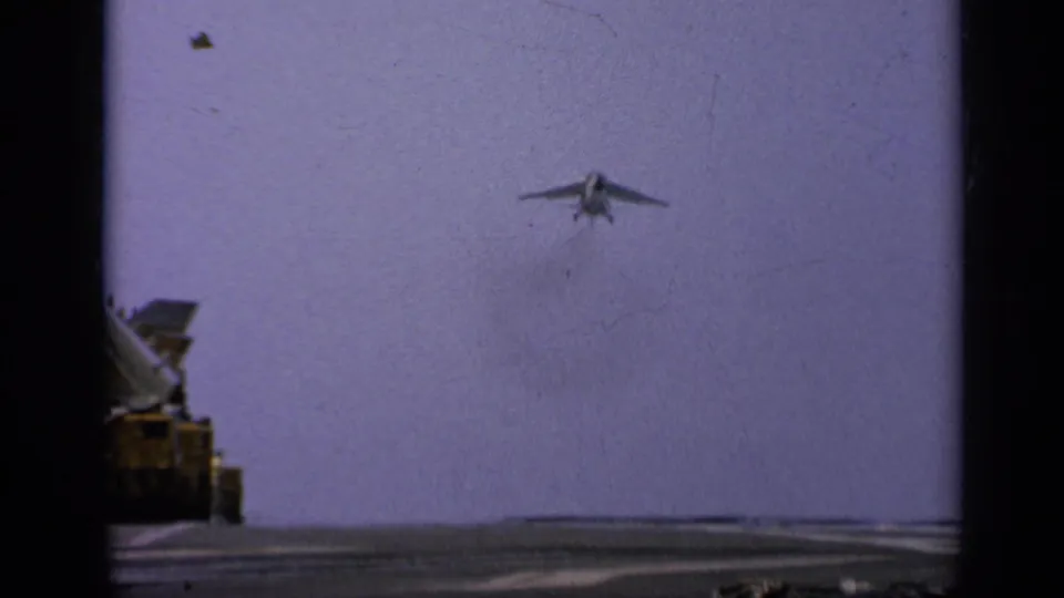 A U.S. Navy jet approaches the carrier deck in 1962 Italy footage, with deck machinery visible at the edge.