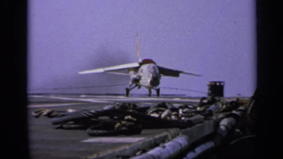 A U.S. Navy jet is stopped or slowing on the carrier deck, with a yellow-shirted deck crew member nearby in the 1962 Italy footage.