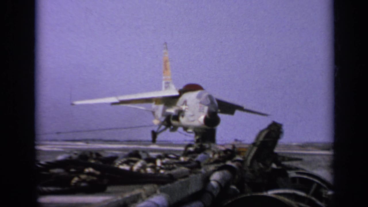 1962 Italy-labeled 8mm frame showing a U.S.-marked jet on an aircraft carrier deck with equipment in front.