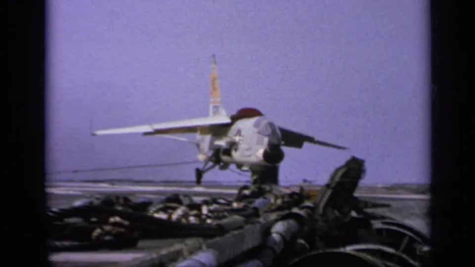 1962 Italy-labeled 8mm frame showing a U.S.-marked jet on an aircraft carrier deck with equipment in front.