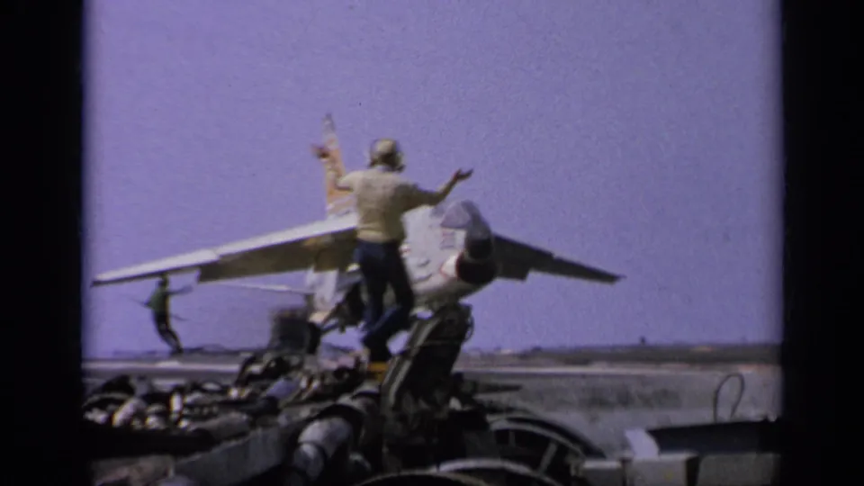 A crew member gestures beside a U.S.-marked jet on an aircraft carrier deck in the supplied 1962 Italy footage.