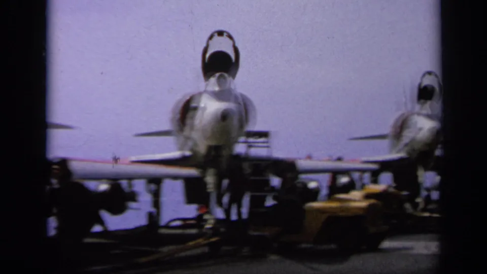 Rear view of two jet aircraft on a carrier deck, with deck equipment and silhouetted crew nearby.