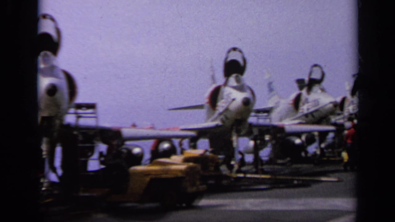 Jet fighters lined up on an aircraft carrier deck with deck equipment nearby.
