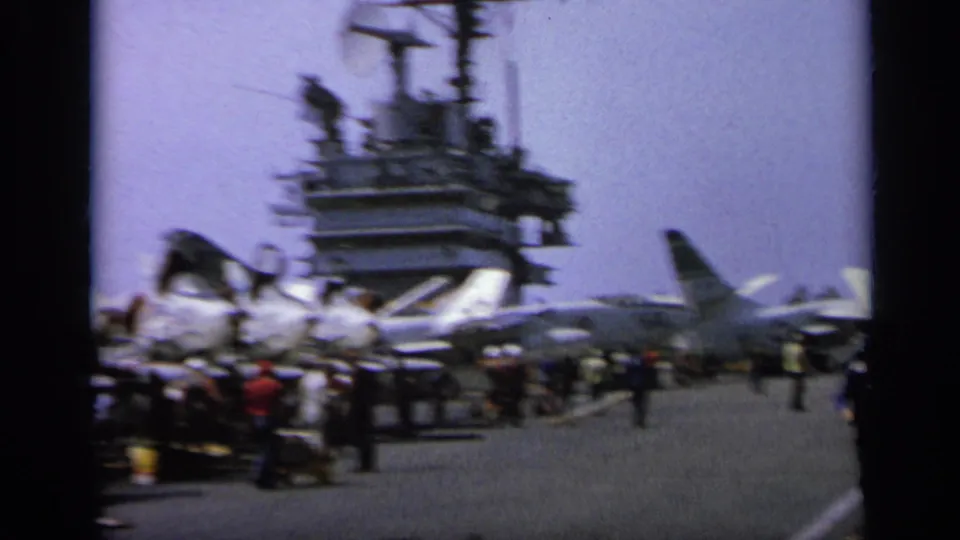 Wide 8mm view of a busy carrier deck in Italy, with jet aircraft, people, carts, and the island structure visible.