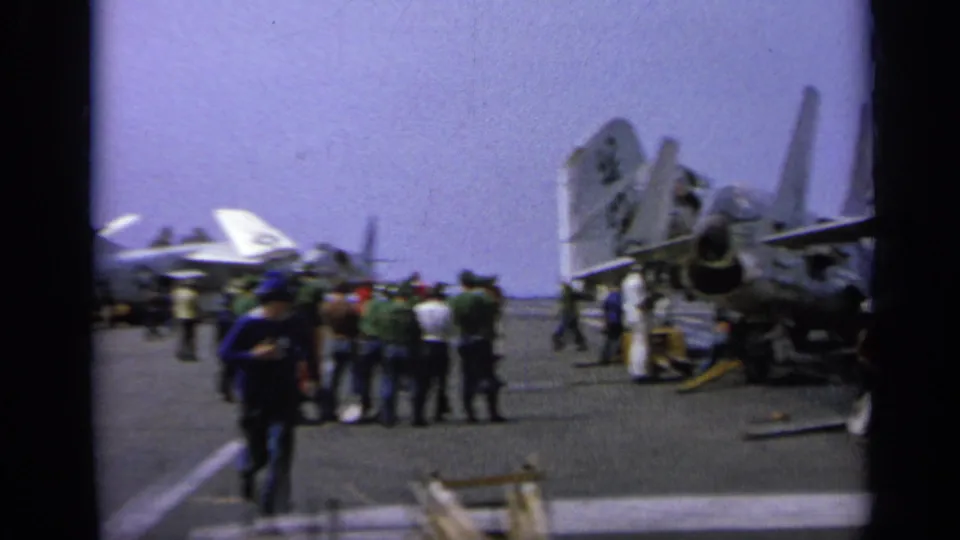 1962 Italy home-movie view of visitors or crew walking between parked jet fighters on an aircraft carrier deck.