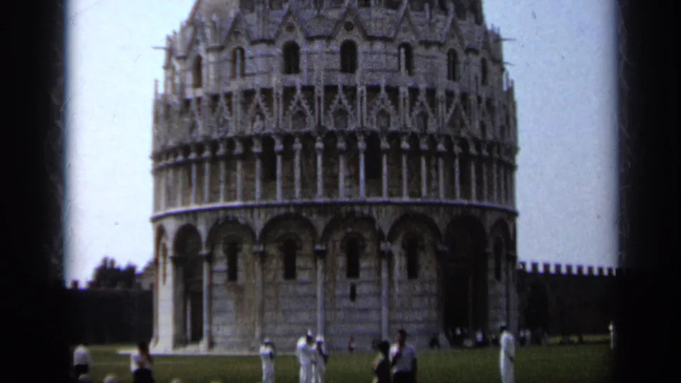 1962 8mm tourist view of the Leaning Tower of Pisa, showing the lower arches, grassy foreground, and visitors near the base.