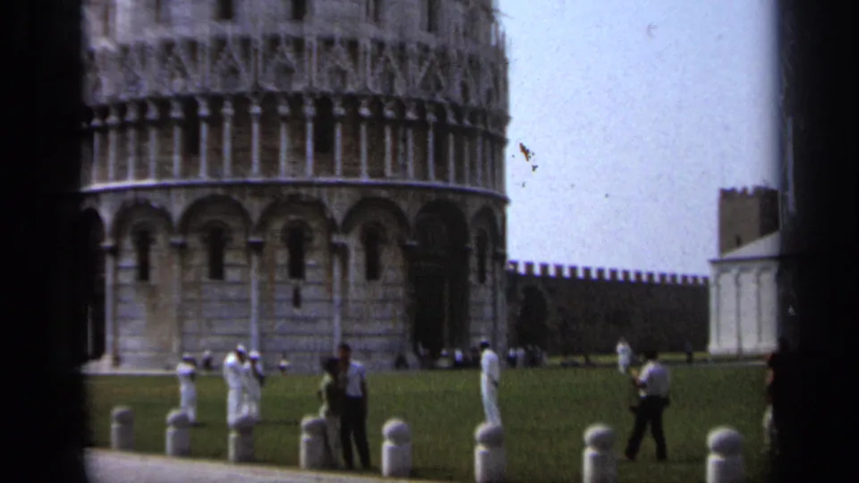 Wider 1962 Pisa view with visitors walking across the grass below the Leaning Tower and a wall in the background.