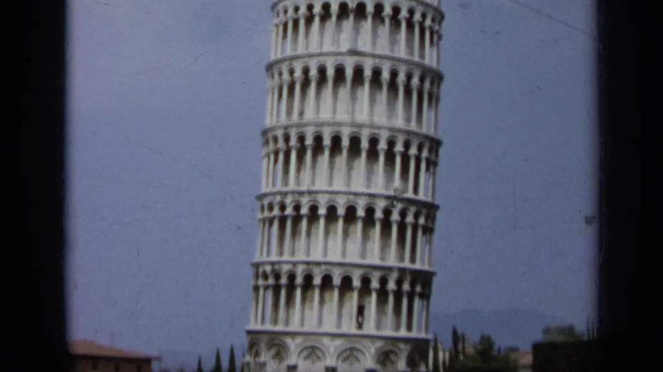 1962 color 8mm frame tightly framing the Leaning Tower of Pisa with sky, trees and a low building at the edge.