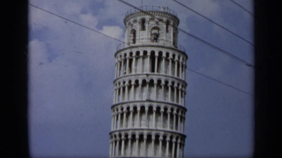 1962 Italy 8mm frame showing a partial tilted view of the Leaning Tower of Pisa, with arcaded levels, clouds, and diagonal overhead wires.