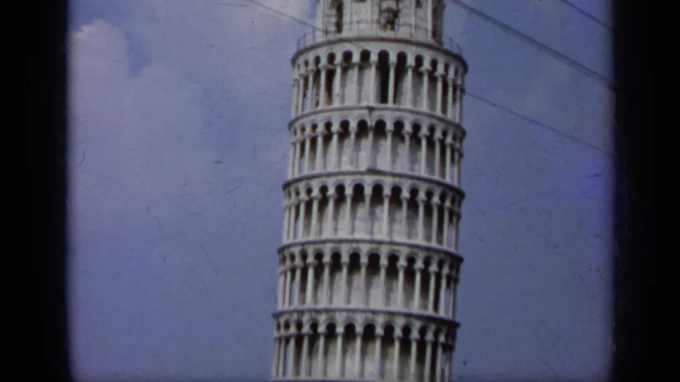 1962 Italy 8mm frame of the Leaning Tower of Pisa cropped at top and base, showing white arcades, blue sky, clouds, and crossing wires.