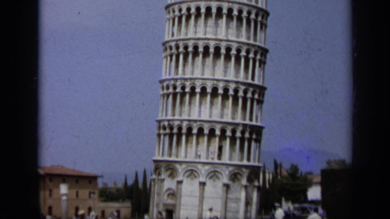 1962 8mm view of the Leaning Tower of Pisa with lower arches, distant building, trees, and landmark grounds.