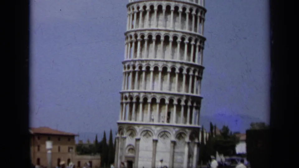1962 8mm view of the Leaning Tower of Pisa with lower arches, distant building, trees, and landmark grounds.