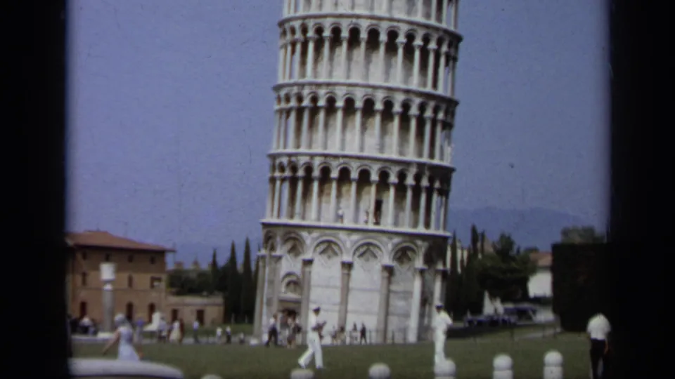 1962 8mm Italy frame shows the Leaning Tower of Pisa, lawn, distant tourists, white posts, and nearby architecture.