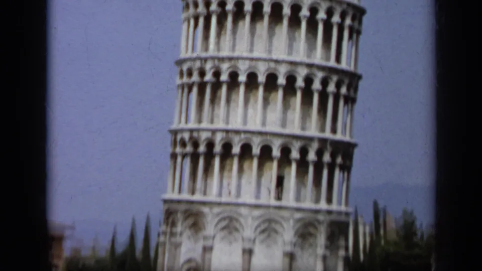 Cropped 1962 8mm view of the Leaning Tower of Pisa base and lower galleries, with trees and sky beyond.